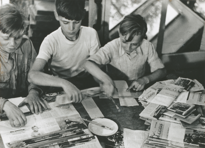 876624 Portret van drie jongens die tijdschriften 'De Stad Gods' aan het vouwen zijn voor verdere distributie, in het ...